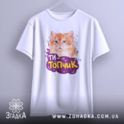 Футболка ти топчик, біла, зображена на вішалці, фон світло-сірий, принт з оранжевим котом та написом 'ТИ-Топчик' у фіолетовій обгортці.