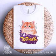 Футболка ти топчик, біла, складена на плетеній основі, фон білий, принт з оранжевим котом та написом 'ТИ-Топчик' у фіолетовій обгортці.