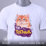 Футболка ти топчик, біла, зображена зверху, фон світло-сірий, принт з оранжевим котом та написом 'ТИ-Топчик', на футболці є етикетка з зеленим шнурком.
