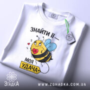 Футболка удача поруч, біла футболка, складена на столі, з принтом жовтої бджоли з блакитними крилами, що тримає червоне серце, з зеленим ярликом.