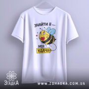 Футболка удача поруч, біла футболка, підвішена на вішалці, з принтом жовтої бджоли з блакитними крилами, що тримає червоне серце, на фоні світло-сірого кольору.