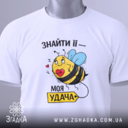 Футболка удача поруч, біла футболка, складена на столі, з принтом жовтої бджоли з блакитними крилами, що тримає червоне серце, з зеленим ярликом.