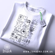 Футболка Україна мій світ, біла, складена на поверхні, фон світлий, принт чорного кольору з малюнками та текстами, на футболці є зелений ярлик.