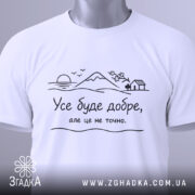 Футболка усе буде добре, на білому фоні, зображена зверху, складена, з етикеткою зеленого кольору, принт з пейзажем та текстом чорного кольору.