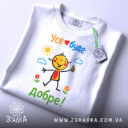 Футболка усе буде добре. Біла футболка, складена на поверхні, з кольоровим принтом усміхненого персонажа, текстом 'Усе❤️буде добре!' та яскравими елементами природи. На футболці є етикетка з зеленим шнурком.