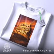 Футболка усім абсолютно пофіг. Біла футболка, зображена з верхнього ракурсу, принт на футболці з написом 'УСІМ АБСОЛЮТНО ПОФІГ', фон світлий, на футболці є етикетка з зеленим шнурком.