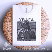 Футболка увага небезпечно, біла, зображена зверху на плетеній основі, фон світлий, принт з зображенням персонажа в чорному кольорі, текст на українській мові.