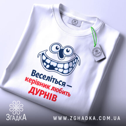 Футболка веселий керівник. Біла футболка, складена на поверхні, з видимим принтом усміхненого персонажа та текстом "Веселіться — керівник любить ДУРНІВ" у синьому та червоному кольорах. На футболці є етикетка з зеленим шнурком.