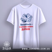 Футболка веселий керівник. Біла футболка, зображена з фронтального ракурсу на світло-сірому фоні. Принт з усміхненим персонажем та текстом "Веселіться — керівник любить ДУРНІВ" у синьому та червоному кольорах.