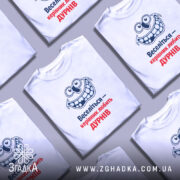 Футболка веселий керівник. Біла футболка, зображена з фронтального ракурсу на манекені, фон світло-сірий. Принт з усміхненим персонажем та текстом "Веселіться — керівник любить ДУРНІВ" у синьому та червоному кольорах.