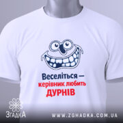Футболка веселий керівник. Біла футболка, складена на поверхні, з видимим принтом усміхненого персонажа та текстом "Веселіться — керівник любить ДУРНІВ" у синьому та червоному кольорах. На футболці є етикетка з зеленим шнурком.