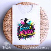 Футболка вільна жінка, білого кольору, зображена зверху на плетеній кошику, фон білий, принт з написом 'Вільна Жінка' у яскравих кольорах.