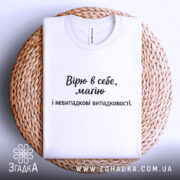Футболка віра в себе, має білий колір, зображена в складеному вигляді на плетеній основі, фон білий, принт чорного кольору з текстом "Вірю в себе, маю і невипадкові випадковості.".