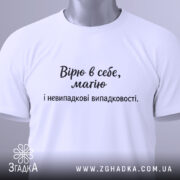 Футболка віра в себе, має білий колір, зображена в складеному вигляді, фон світло-сірий, принт чорного кольору з текстом "Вірю в себе, маю і невипадкові випадковості." на видному місці, з етикеткою зеленого кольору.