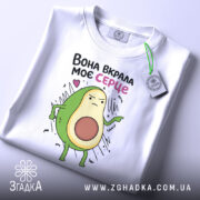 Футболка вкрала моє серце. Біла футболка, складена на столі, з принтом авокадо, що виглядає впевнено. На футболці є етикетка з зеленим шнурком. Фон світлий, з легким відблиском.