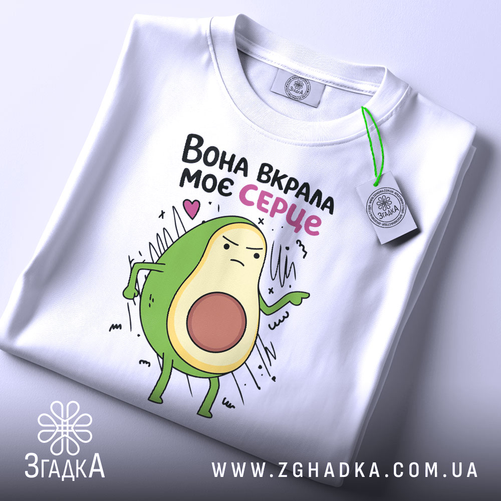 Футболка вкрала моє серце. Біла футболка, складена на столі, з принтом авокадо, що виглядає впевнено. На футболці є етикетка з зеленим шнурком. Фон світлий, з легким відблиском.