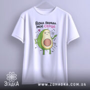 Футболка вкрала моє серце. Біла футболка, підвішена на вішалці, з принтом авокадо, що показує пальцем. Фон світлий, з легким градієнтом. Принт яскравий, з веселими деталями.