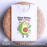 Футболка вкрала моє серце. Біла футболка, складена на плетеному кошику, з принтом авокадо, що має серйозний вираз обличчя. Фон світлий, з текстурою плетеного кошика. Принт яскравий, з кольоровими деталями.