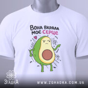 Футболка вкрала моє серце. Біла футболка, складена на столі, з принтом авокадо, що виглядає впевнено. На футболці є етикетка з зеленим шнурком. Фон світлий, з легким відблиском.