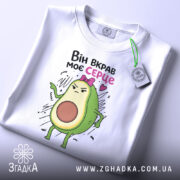 Футболка вкрав моє серце. Біла футболка, складена на поверхні, з принтом авокадо, яке виглядає сердито. Принт займає центральну частину, а на комірці видно етикетку. Фон світлий, простий.