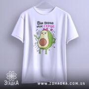 Футболка вкрав моє серце. Біла футболка, підвішена на вішалці, з принтом авокадо, яке має сердитий вираз. Принт яскравий, розташований на грудях, фон світлий, без зайвих елементів.