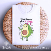 Футболка вкрав моє серце. Біла футболка, складена на плетеній кошику, з принтом авокадо, яке виглядає сердито. Принт яскравий, фон натуральний, з текстурою кошика.