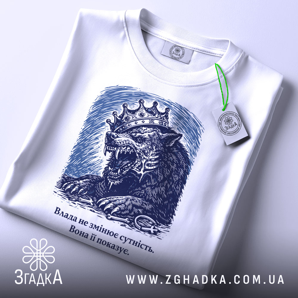 Футболка влада і сутність, білого кольору, зображена зверху, фон світло-сірий, принт з малюнком лева з короною в темно-синіх тонах, текст під малюнком, на футболці є етикетка з зеленим шнурком.