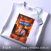 Футболка все горить. Біла футболка, складена на поверхні, з принтом, що зображує чоловіка за комп'ютером в офісі, оточеного вогнем. Текст "Все горить. Мені по*уй" виконаний у яскравих кольорах.