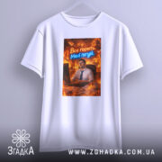 Футболка все горить. Біла футболка, підвішена на вішалці, з принтом, на якому зображений чоловік за комп'ютером в офісі, оточений вогнем. Текст "Все горить. Мені по*уй" яскраво виділяється.