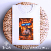 Футболка все горить. Біла футболка, складена на плетеній основі, з принтом, що зображує чоловіка за комп'ютером в офісі, оточеного вогнем. Текст "Все горить. Мені по*уй" яскраво контрастує з фоном.