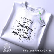Футболка всесвіт на боці, біла, складена на поверхні, з видимим етикеткою, фон світло-сірий, принт чорний з написом 'всесвіт завжди на боці мрійників' та декоративними зірочками.