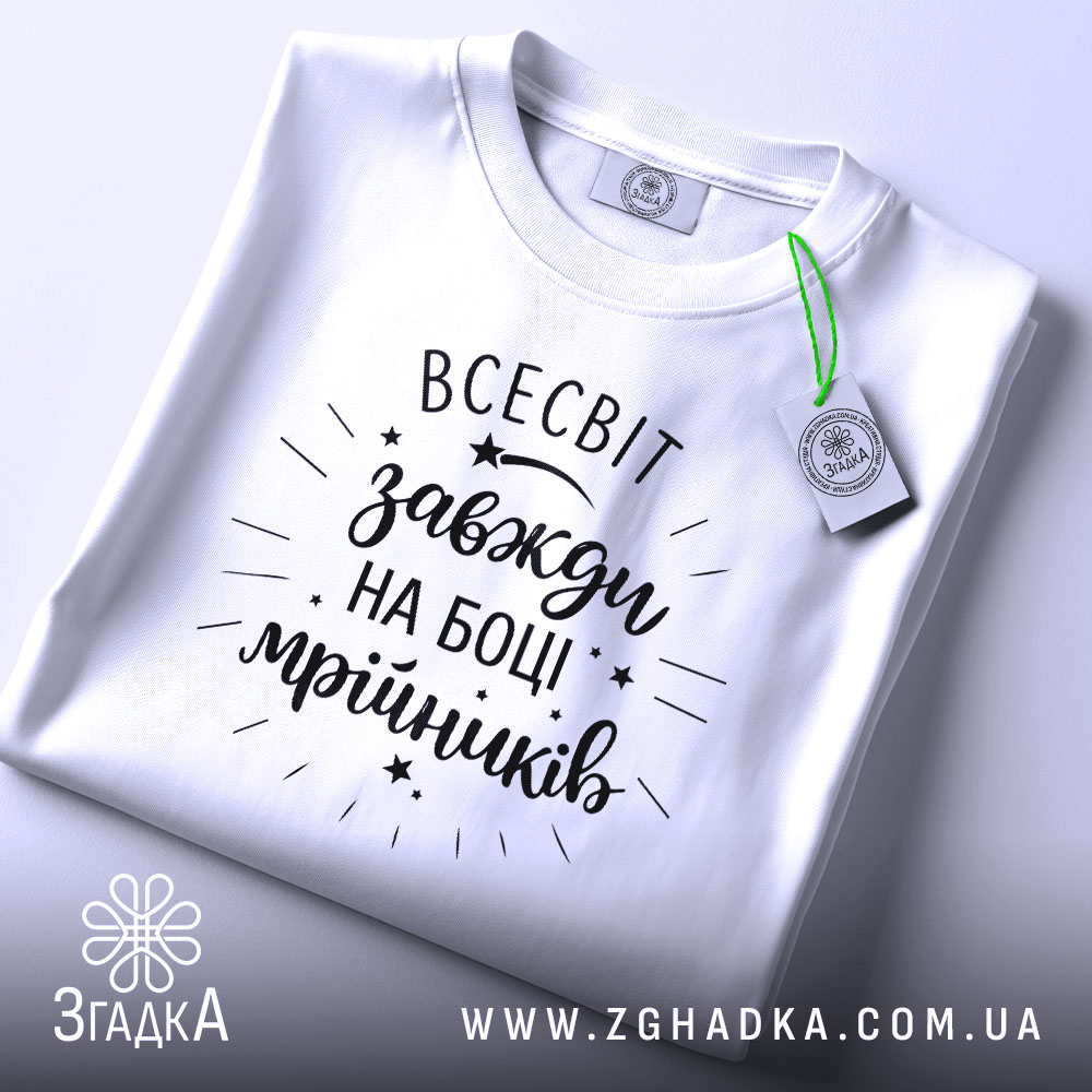 Футболка всесвіт на боці, біла, складена на поверхні, з видимим етикеткою, фон світло-сірий, принт чорний з написом 'всесвіт завжди на боці мрійників' та декоративними зірочками.