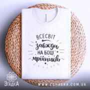 Футболка всесвіт на боці, біла, складена на плетеній кошику, фон світло-сірий, принт чорний з написом 'всесвіт завжди на боці мрійників' та декоративними зірочками.