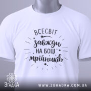 Футболка всесвіт на боці, біла, складена на поверхні, з видимим етикеткою, фон світло-сірий, принт чорний з написом 'всесвіт завжди на боці мрійників' та декоративними зірочками.