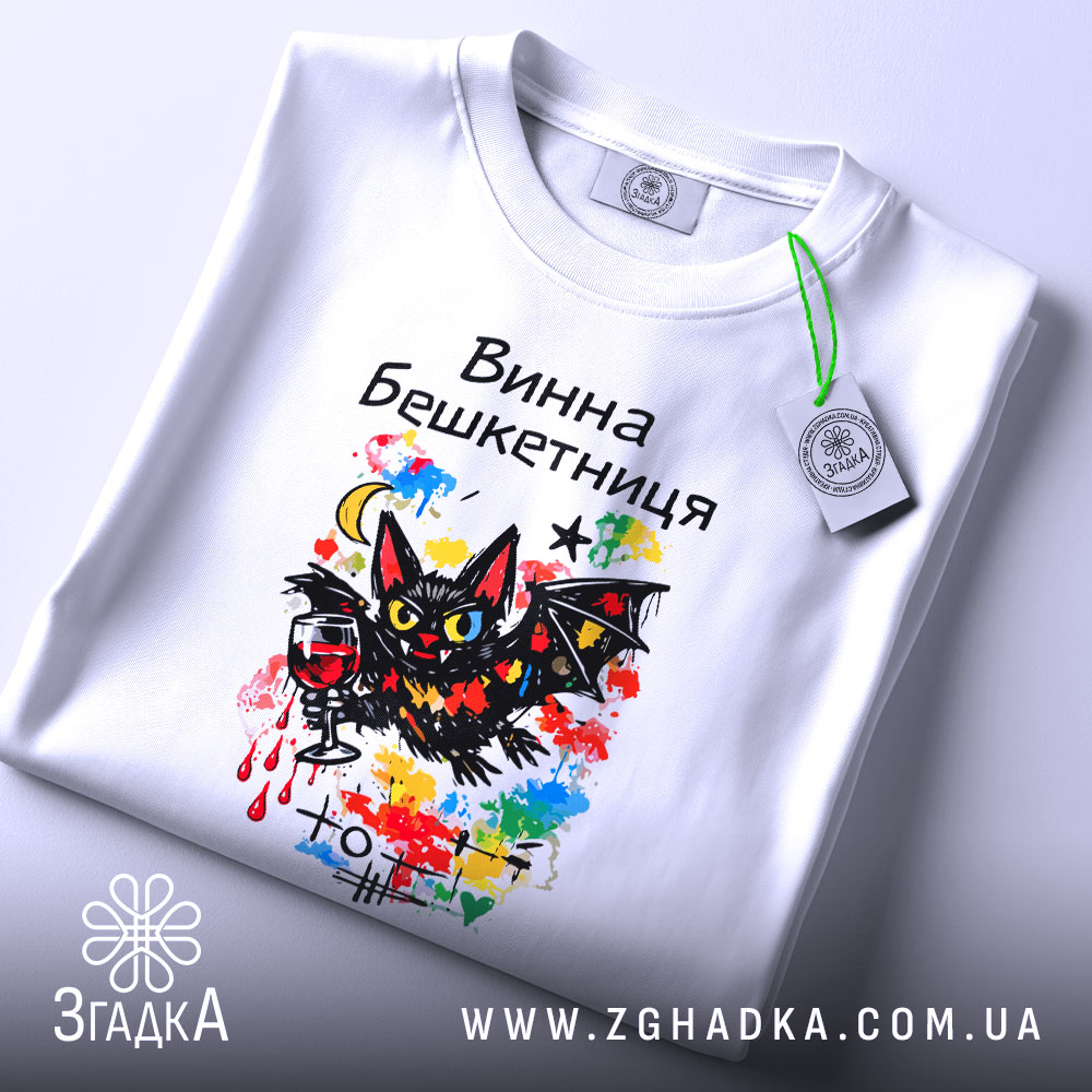 Футболка винна бешкетниця, білого кольору, складена на поверхні, фон світлий, принт з чорним котом, червоним вином та яскравими кольоровими акцентами, на футболці є етикетка.