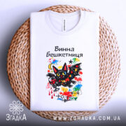 Футболка винна бешкетниця, білого кольору, складена на плетеній кошику, фон світлий, принт з чорним котом, червоним вином та яскравими кольоровими елементами, виглядає затишно.