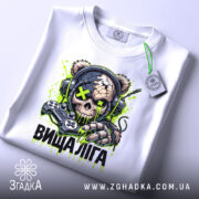 Футболка вища ліга. Біла футболка, розкладена на поверхні, з яскравим принтом черепоподібної ведмедиці в навушниках, що тримає геймпад. Принт має зелені та чорні деталі, а на футболці висить етикетка. Фон світлий.
