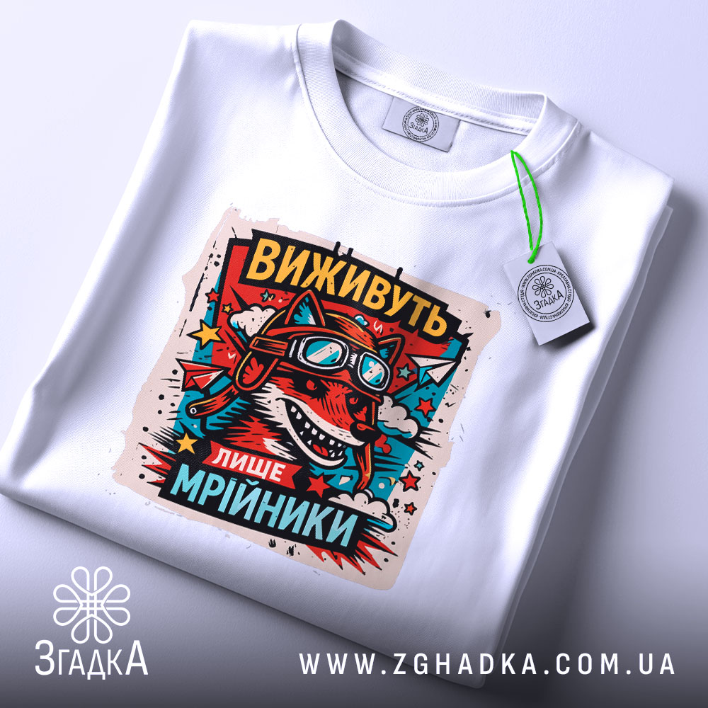 Футболка виживуть лише мрійники. Біла футболка, складена на столі, з принтом червоного вовка в окулярах та шоломі, оточеного кольоровими зірками. На футболці є етикетка з зеленим шнурком. Фон світло-сірий.
