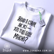 Футболка з філософським написом, білого кольору, складена на поверхні, фон світло-сірий, принт чорного кольору з текстом: 'Якщо я стану, як усі — хто тоді буде мною?', на комірці видно етикетку.