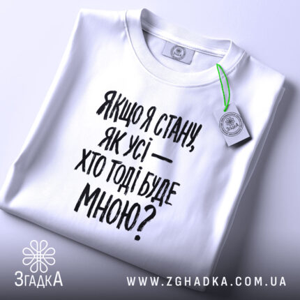 Футболка з філософським написом, білого кольору, складена на поверхні, фон світло-сірий, принт чорного кольору з текстом: 'Якщо я стану, як усі — хто тоді буде мною?', на комірці видно етикетку.