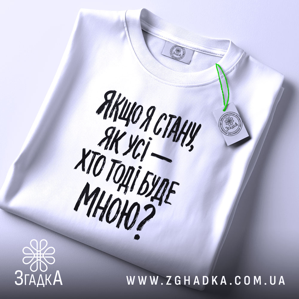 Футболка з філософським написом, білого кольору, складена на поверхні, фон світло-сірий, принт чорного кольору з текстом: 'Якщо я стану, як усі — хто тоді буде мною?', на комірці видно етикетку.