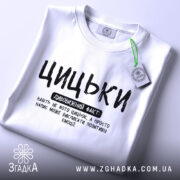 Футболка з написом цицьки, білого кольору, складена на поверхні з нейтральним фоном, принт чорного кольору з великими буквами, текст розташований по центру, з етикеткою на комірці.