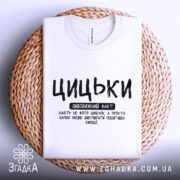 Футболка з написом цицьки, білого кольору, складена на плетеній основі з нейтральним фоном, принт чорного кольору з великими буквами, текст розташований по центру.