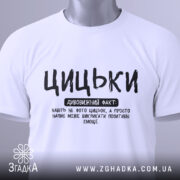 Футболка з написом цицьки, білого кольору, складена на поверхні з нейтральним фоном, принт чорного кольору з великими буквами, текст розташований по центру, з етикеткою на комірці.