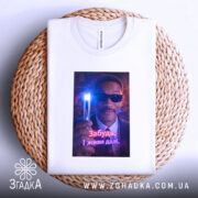 Футболка забудь і живи. Біла футболка, складена на плетеній основі, з яскравим принтом чоловіка в окулярах, що тримає світловий прилад. Принт виділяється на фоні текстури.