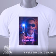 Футболка забудь і живи. Біла футболка, складена на поверхні, з яскравим принтом чоловіка в окулярах, що тримає світловий прилад. На футболці є етикетка з зеленим шнурком.