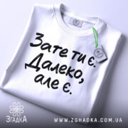 Футболка зате ти є. Білий колір, зображена зверху, складена на поверхні, фон світлий, принт чорного кольору з текстом 'Зате ти є. Далеко, але є.', з етикеткою зеленого кольору.