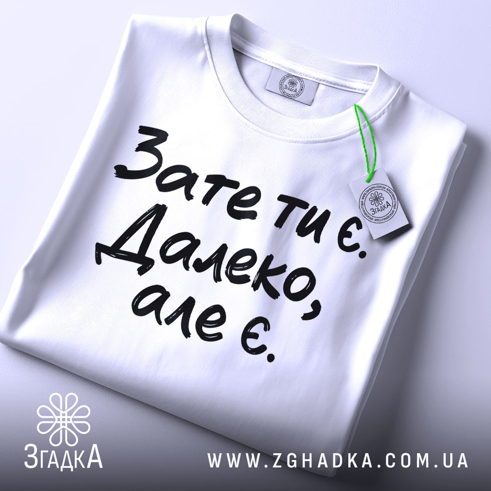 Футболка зате ти є. Білий колір, зображена зверху, складена на поверхні, фон світлий, принт чорного кольору з текстом 'Зате ти є. Далеко, але є.', з етикеткою зеленого кольору.