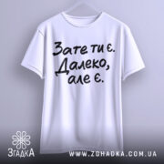 Футболка зате ти є. Білий колір, зображена на вішалці, фон світлий, принт чорного кольору з текстом 'Зате ти є. Далеко, але є.'