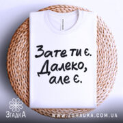 Футболка зате ти є. Білий колір, зображена зверху, складена на плетеній основі, фон світлий, принт чорного кольору з текстом 'Зате ти є. Далеко, але є.'